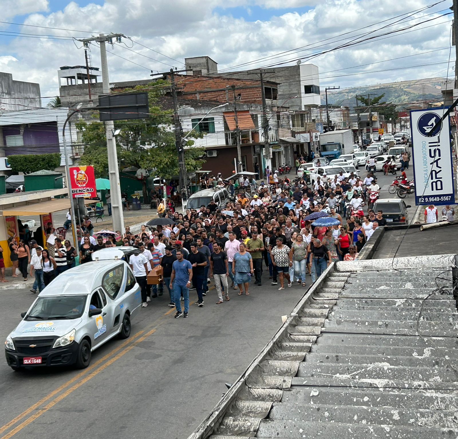 Vista aérea de uma rua movimentada em União dos Palmares, onde um carro funerário prata lidera um grande cortejo a pé. Uma multidão de pessoas segue o veículo, ocupando toda a extensão da via entre prédios comerciais e residenciais sob um céu nublado.