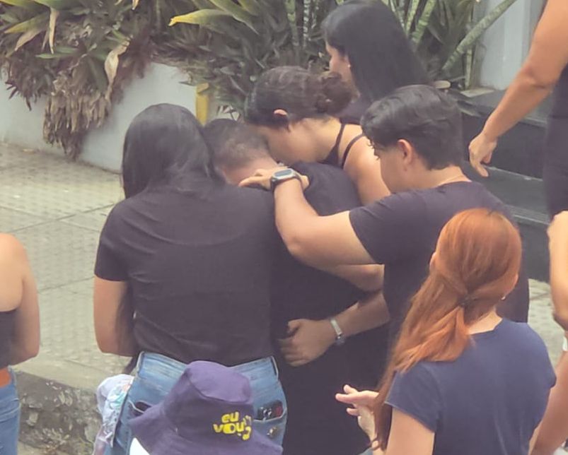O influenciador Murilo Dias, visto de costas vestindo uma camiseta preta, sendo amparado e abraçado por três amigos durante o cortejo fúnebre de sua irmã em União dos Palmares.