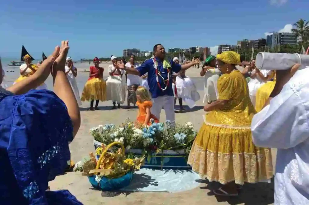 Pessoas vestidas com roupas típicas de religiões de matriz africana realizando celebração de Iemanjá na praia, com flores e oferendas na areia, em Maceió.