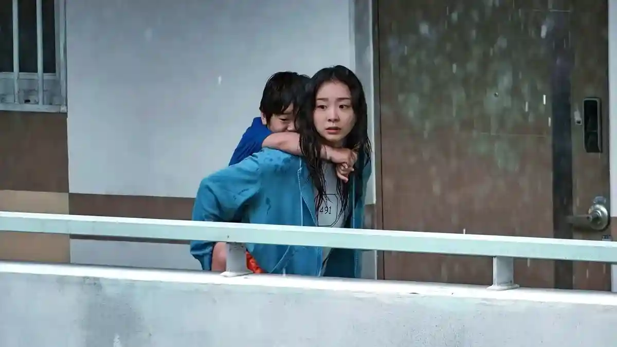 mulher carrega crianca nas costas durante chuva intensa tentando escapar de enchente em cena do filme a grande inundacao da netflix