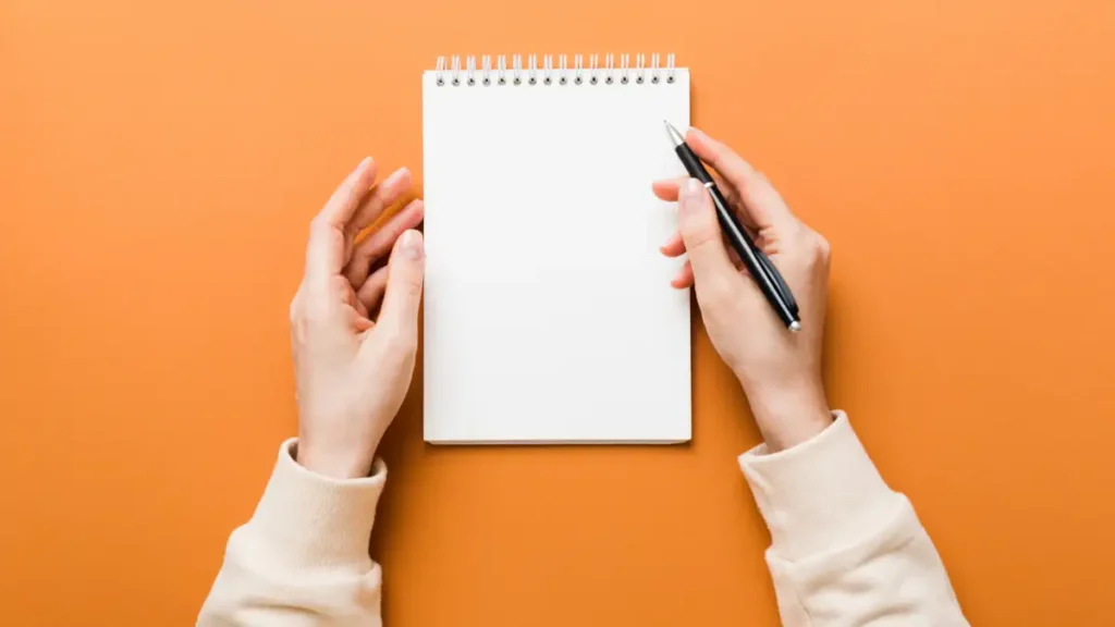 pessoa segurando caneta e caderno em branco sobre fundo laranja em ambiente de planejamento e anotacoes