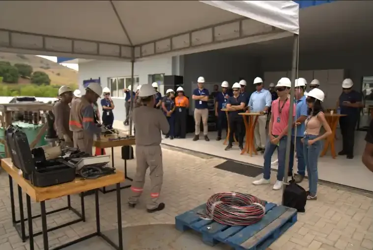 Sob uma tenda branca em área externa, um grupo de profissionais da imprensa e da Equatorial Alagoas, todos usando capacetes brancos, observa uma demonstração técnica. Em primeiro plano, uma mesa de madeira exibe equipamentos de segurança e, no chão, há um rolo de cabos elétricos sobre um palete azul. Técnicos uniformizados manuseiam ferramentas em uma bancada ao lado.
