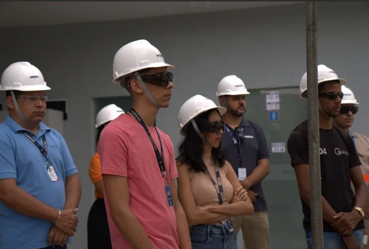Em primeiro plano, um jovem de camiseta rosa e óculos de proteção escuros observa atentamente uma demonstração. Ao seu lado, uma mulher de blusa bege sem mangas e óculos escuros também presta atenção. Ambos usam capacetes brancos de segurança com o logotipo da Equatorial. Outros profissionais e técnicos uniformizados aparecem ao fundo, em um ambiente externo.
