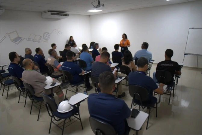 Sala de treinamento com cerca de 15 pessoas sentadas em cadeiras universitárias, assistindo a uma palestra. Ao fundo, uma palestrante vestindo uma camisa polo laranja da Equatorial fala em pé em frente a uma parede branca. Na parede lateral esquerda, há desenhos de traço fino representando um notebook, um livro aberto e um globo terrestre.