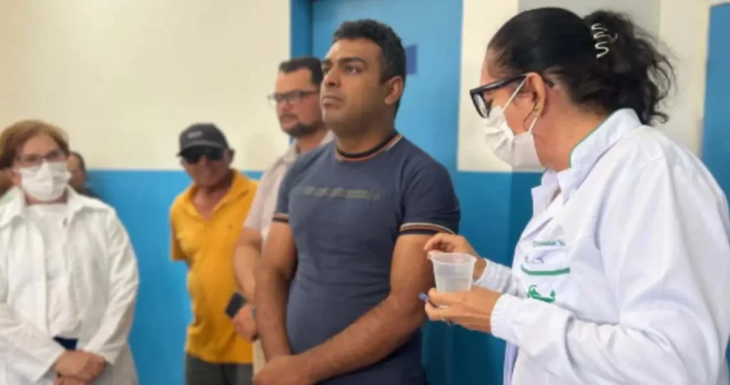 Uma profissional de saúde, de jaleco branco e máscara, segura um copo com água enquanto orienta um homem de camiseta azul marinho e braços cruzados. Ao fundo, outros moradores e uma profissional de branco acompanham a palestra em um ambiente interno com paredes azuis e brancas.