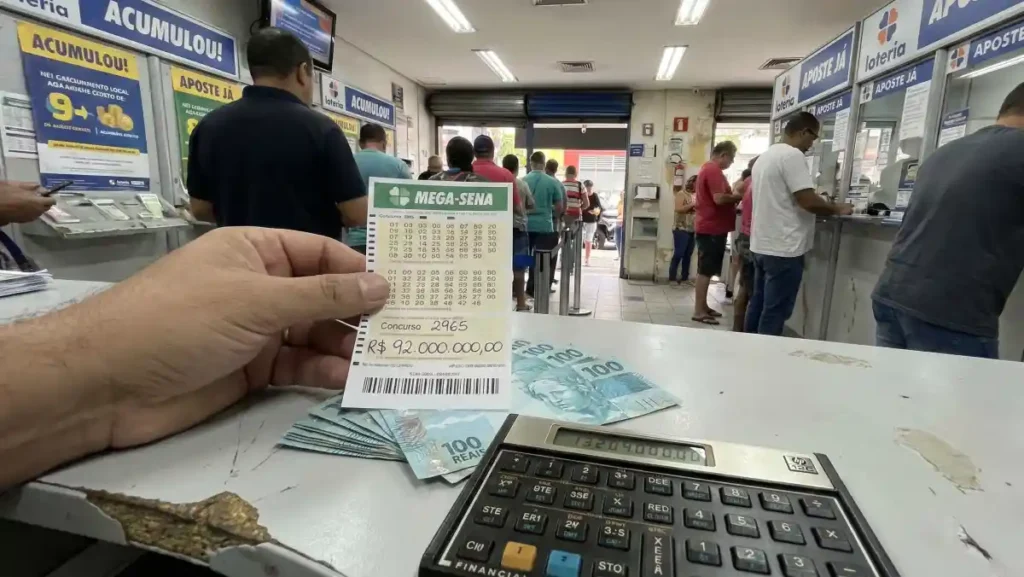 apostador segura volante da Mega-Sena em lotérica com cédulas de real e calculadora sobre o balcão e fila de atendimento ao fundo