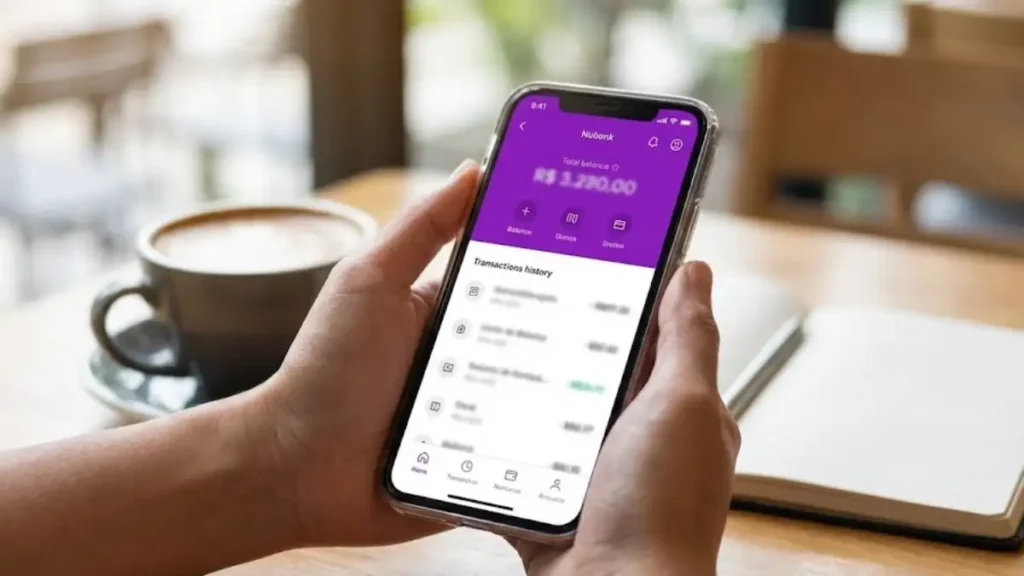 Pessoa segura celular com aplicativo do Nubank aberto sobre mesa de café, em imagem ilustrativa de conta digital e transações
