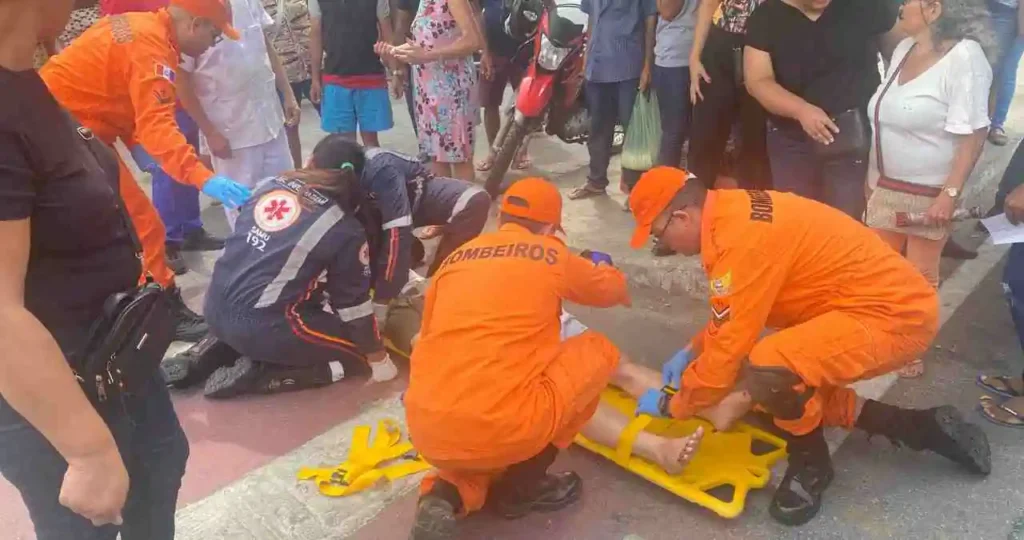 Cena de um resgate em via pública. Três bombeiros vestindo uniformes laranja e uma socorrista do SAMU com macacão azul marinho prestam atendimento a uma pessoa deitada em uma prancha rígida amarela no asfalto. Vários populares observam o socorro ao redor. Uma motocicleta vermelha está estacionada próxima ao grupo.