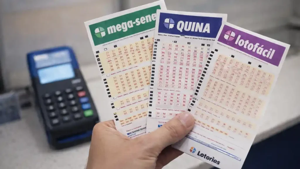Mão segura cartelas de loteria da Mega-Sena, Quina e Lotofácil ao lado de terminal de apostas, em casa lotérica
