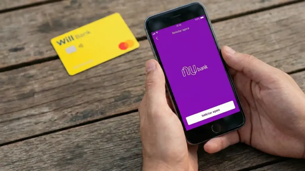 mãos seguram celular com aplicativo do Nubank aberto sobre mesa de madeira enquanto cartão do Will Bank aparece ao fundo