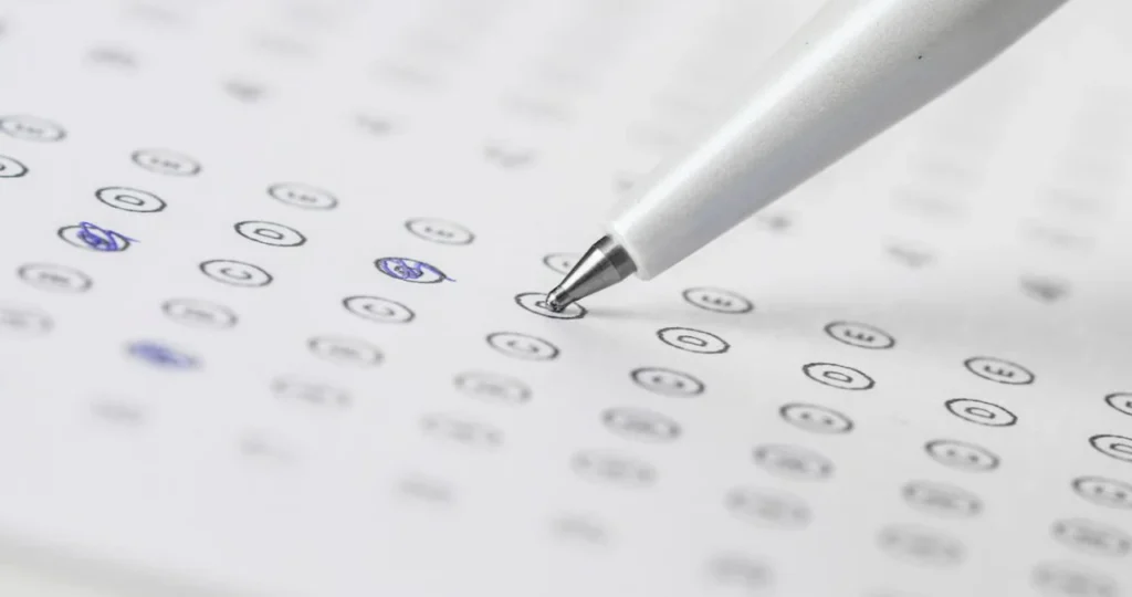 Close-up em ângulo diagonal de uma caneta prateada com ponta esférica tocando um cartão de respostas de concurso público. O papel branco exibe colunas de círculos numerados de "A" a "E", com algumas marcações feitas em caneta azul. O fundo é branco e está levemente desfocado.