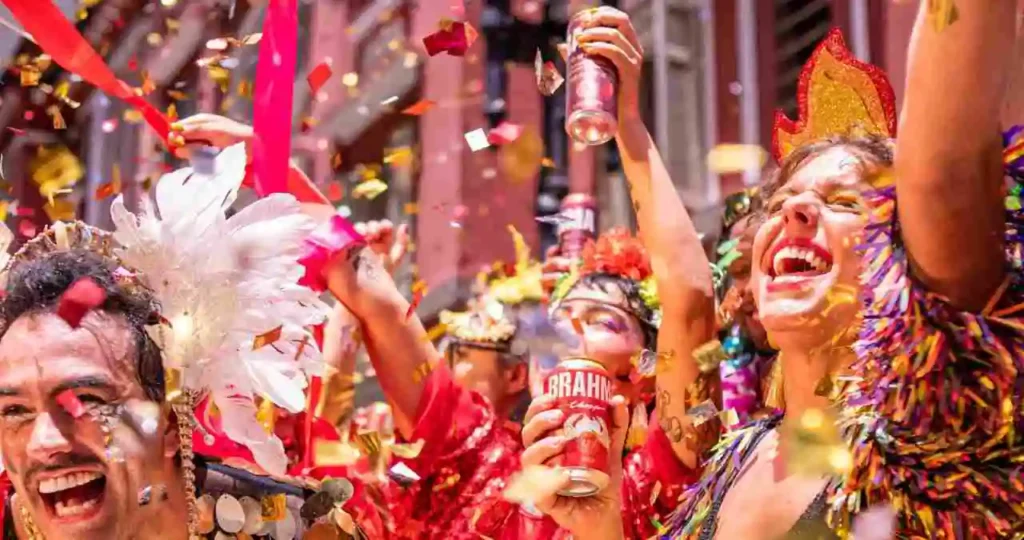 Foliões alegres celebram o Carnaval em meio a uma chuva de confetes dourados. Em destaque, um homem com adereço de penas brancas e uma mulher com tiara de fogo e roupa colorida sorriem enquanto seguram latas de cerveja Brahma.