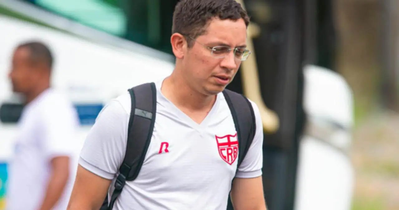 Johanisson Lima (Joba), um homem jovem de óculos e pele clara, veste uma camisa branca de treino com o escudo do CRB no peito e carrega uma mochila preta. Ele caminha em um ambiente externo com um ônibus ao fundo. Johanisson Lima (Joba), um homem jovem de óculos e pele clara, veste uma camisa branca de treino com o escudo do CRB no peito e carrega uma mochila preta. Ele caminha em um ambiente externo com um ônibus ao fundo.