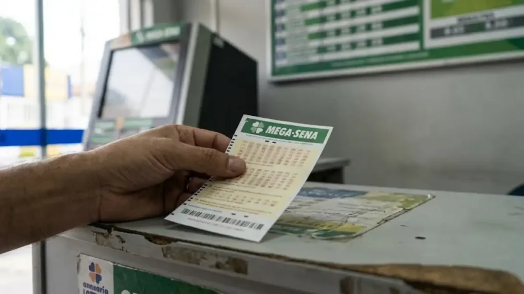 mão segura volante da Mega-Sena da Caixa sobre balcão de lotérica com terminal de apostas ao fundo