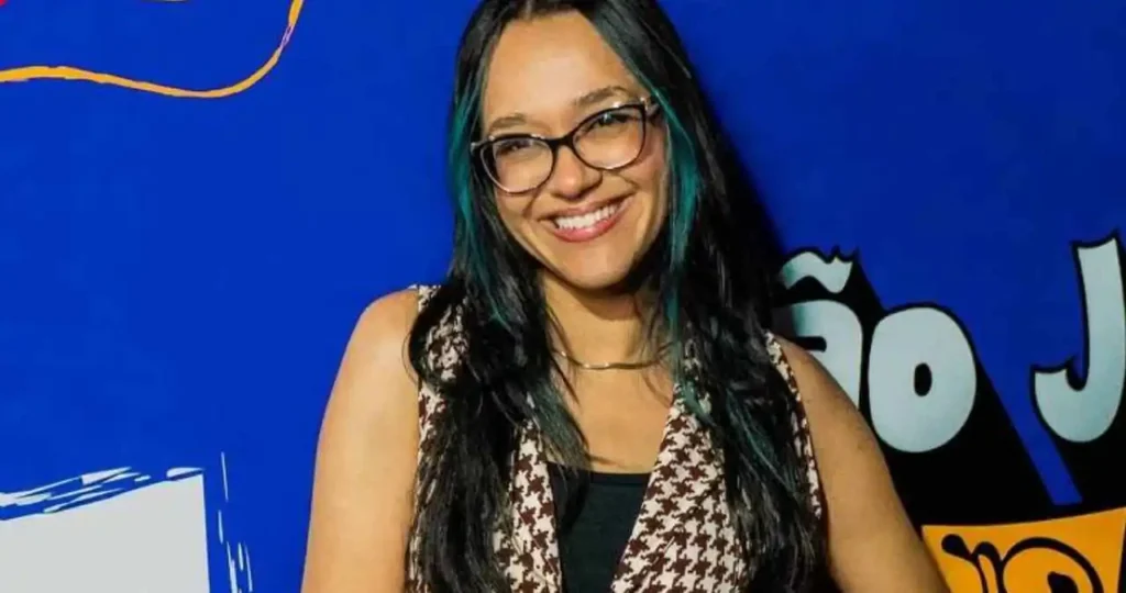 Rafa Jaqueira, uma jovem de pele clara, cabelos longos e escuros com mechas frontais azuis. Ela usa óculos de grau com armação preta, sorri abertamente e veste um colete estampado em tons de marrom e bege sobre uma blusa preta. O fundo é azul vibrante com elementos gráficos em amarelo e vermelho.