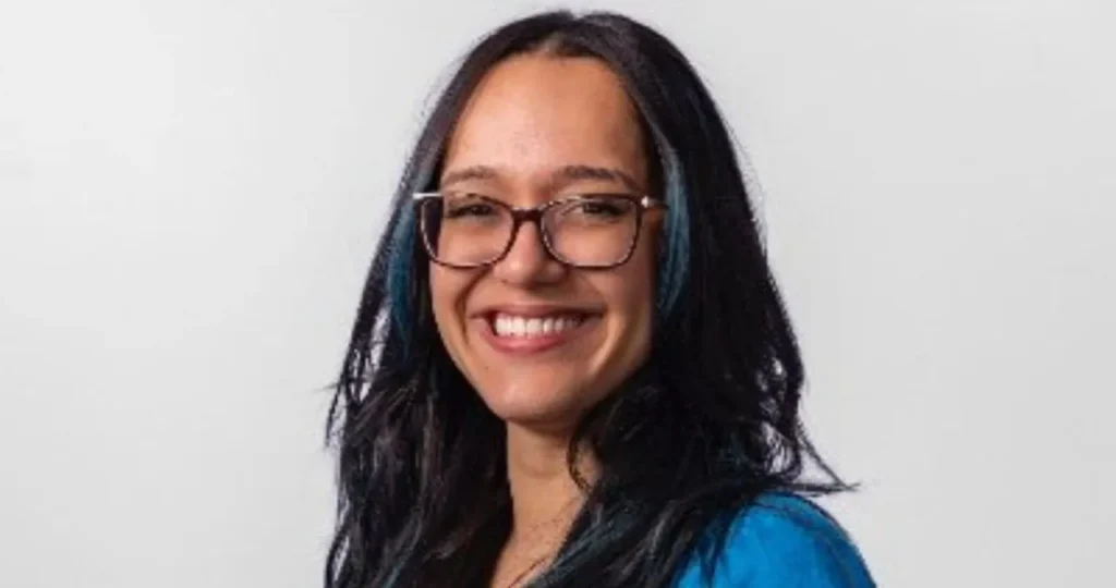 Retrato de Rafa Jaqueira, uma mulher jovem com óculos de armação escura e cabelos longos e pretos com mechas azuis. Ela veste um blazer azul vibrante sobre uma blusa preta e sorri para a foto em um fundo branco.