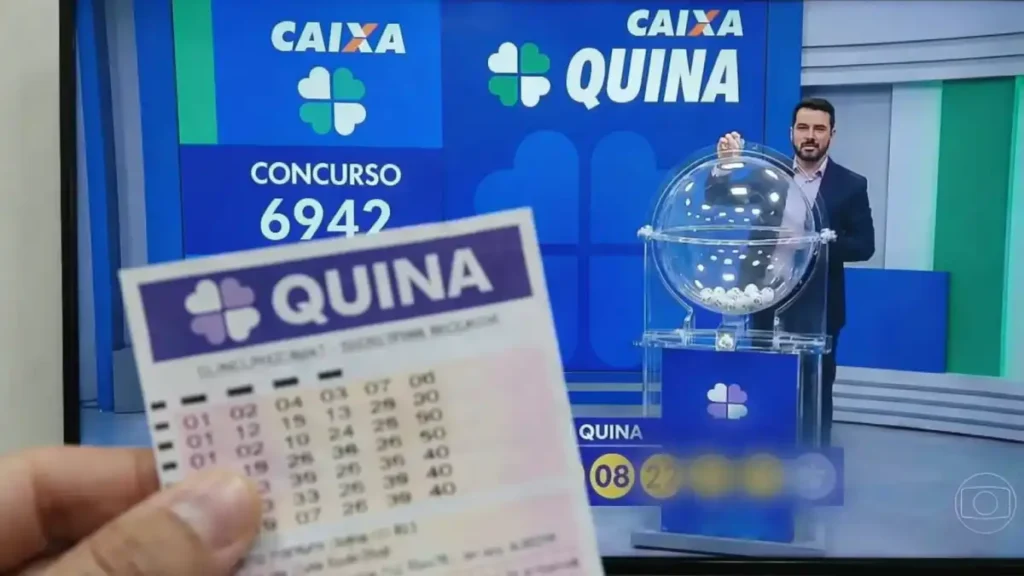 Volante da Quina em primeiro plano com transmissão do sorteio da Caixa ao fundo indicando concurso 6942 e resultado na TV