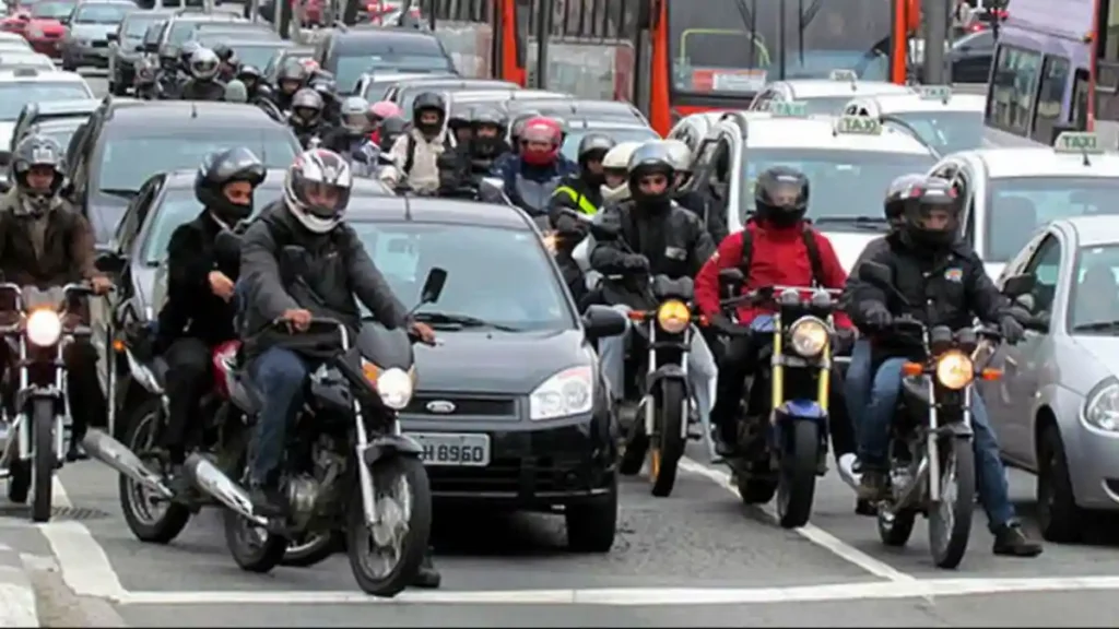 Motociclistas e carros em trânsito intenso em avenida urbana, com ônibus e táxis ao fundo, ilustrando frota de veículos em Alagoas