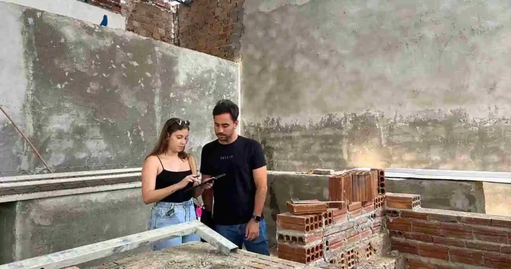 O prefeito de Santana do Mundaú, André Castro, vestindo camiseta preta e calça jeans, observa um tablet segurado por uma engenheira em um canteiro de obras. Ao redor deles, estruturas de alvenaria com tijolos cerâmicos aparentes e vigas de concreto em construção sob um telhado de estrutura metálica.