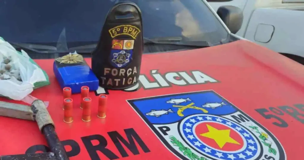 Sobre o capô vermelho de uma viatura da Polícia Militar de Alagoas, com o brasão da corporação em destaque, estão expostos os materiais apreendidos: duas armas de fogo artesanais de cano longo, seis munições de calibre 12 vermelhas, um tablete de maconha envolto em plástico azul e uma sacola com porções menores da droga. Um braçal de couro preto com a inscrição "FORÇA TÁTICA" e o escudo do 5º BPM está posicionado ao centro.