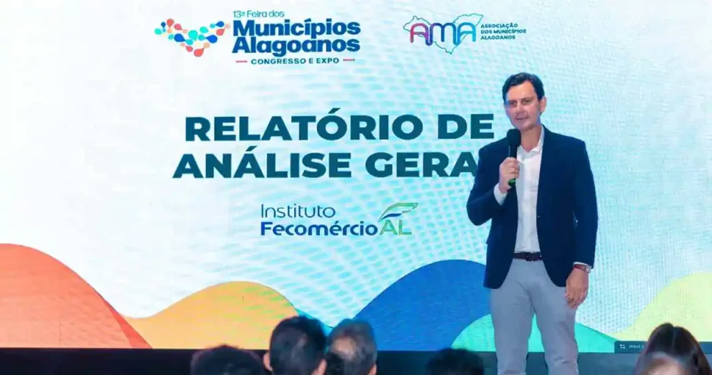 Marcelo Beltrão, um homem de pele clara e cabelos escuros, vestindo um blazer azul marinho sobre uma camisa branca e calças claras, fala ao microfone em um palco. Ao fundo, um telão exibe o título "RELATÓRIO DE ANÁLISE GERAL" com os logotipos da 13ª Feira dos Municípios Alagoanos, AMA e Instituto Fecomércio AL.