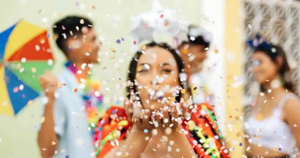 Em primeiro plano, uma mulher com as mãos em concha sopra confetes coloridos em direção à câmera. Ela veste uma roupa colorida de Carnaval e usa um adereço de estrelas brancas na cabeça. Ao fundo, de forma desfocada, outras pessoas celebram, incluindo um homem segurando uma sombrinha de frevo colorida.