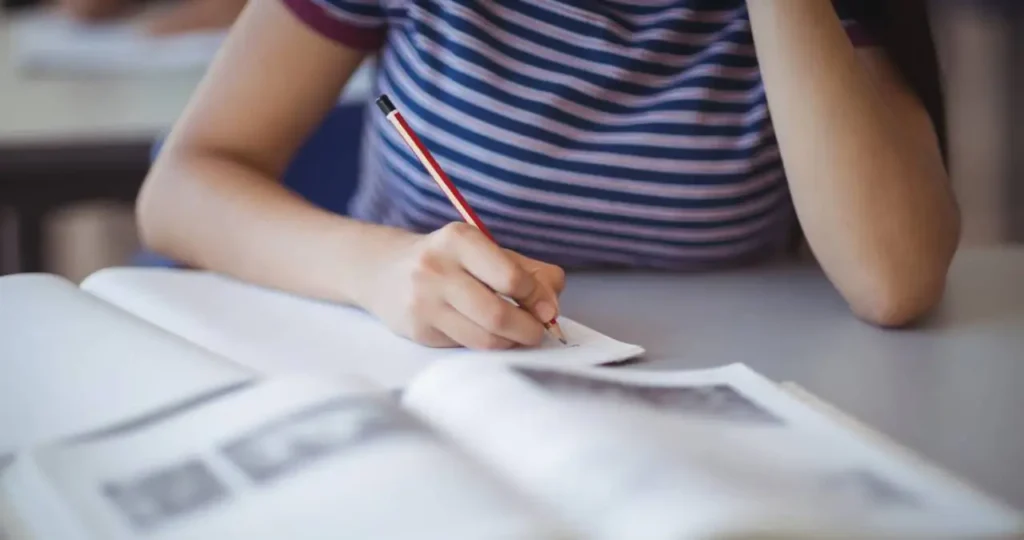 Close-up das mãos de uma estudante vestindo uma blusa listrada de azul e branco, escrevendo com um lápis em um caderno aberto. Em primeiro plano, um livro didático está desfocado sobre a mesa escolar.