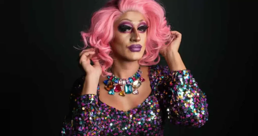 Drag queen com peruca rosa volumosa e vestido de lantejoulas roxas.