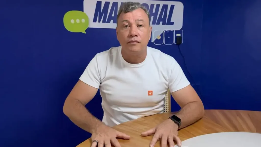 Ex-prefeito de Marechal Deodoro, Cristiano Matheus, sentado à mesa em gravação para redes sociais, com fundo azul
