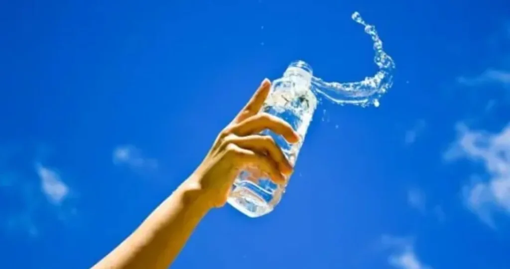 O braço de uma pessoa erguido contra um céu azul vibrante, segurando uma garrafa de água transparente. A água espirra para fora da garrafa, criando uma curva dinâmica no ar. O sol ilumina a cena, destacando o movimento do líquido.