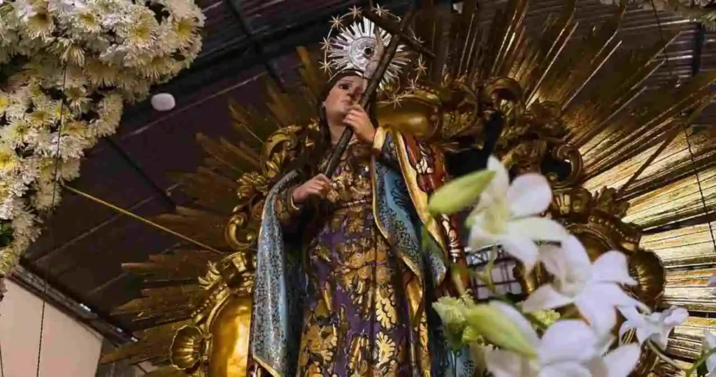Estátua de Santa Maria Madalena em um altar ricamente decorado. A santa veste um manto azul e dourado e segura uma cruz. O altar possui um resplendor dourado ao fundo e está emoldurado por um arco de flores brancas e amarelas. Em primeiro plano, flores brancas e verdes desfocadas.