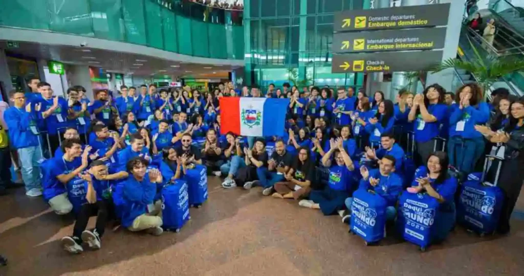 No saguão de um aeroporto, dezenas de estudantes vestindo moletons azuis com a inscrição "Daqui pro Mundo 2025" celebram e aplaudem. Ao centro, um jovem segura a bandeira de Alagoas. À frente, malas de viagem azuis personalizadas estão organizadas. Ao fundo, placas indicam "Embarque doméstico" e "Check-in".