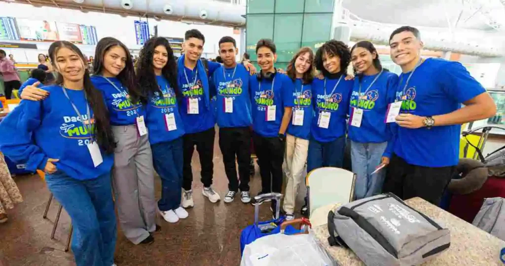Em um saguão de aeroporto moderno, dez jovens (cinco rapazes e cinco moças) sorriem abraçados para a foto. Eles vestem moletons azuis idênticos com o logotipo "Daqui pro Mundo 2025/2026" e usam crachás brancos e roxos. Ao fundo, painéis de voos e outros passageiros são visíveis.