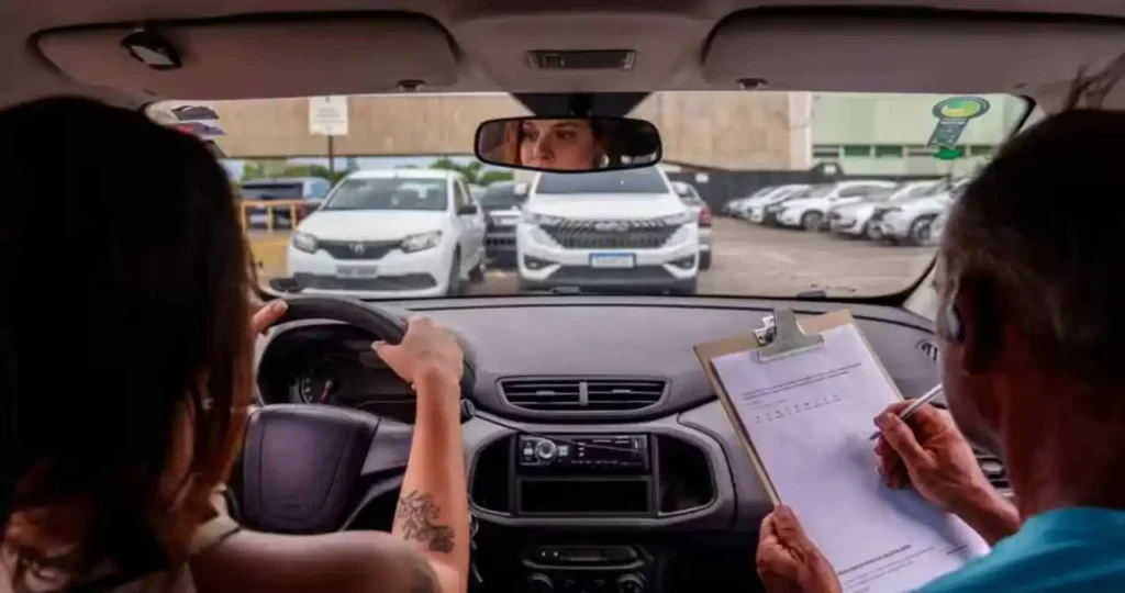Vista do interior de um carro durante um exame de direção. Uma mulher jovem com tatuagem no braço segura o volante, enquanto um examinador ao lado, em uma camiseta azul, escreve em uma prancheta com um formulário de avaliação. Pelo retrovisor interno, é possível ver os olhos da condutora. O carro está em um pátio com outros veículos estacionados ao fundo.