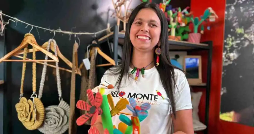 Uma mulher sorridente com cabelos pretos longos segura uma escultura de madeira colorida em formato de cacto com flores vermelhas. Ela veste uma camiseta branca com a estampa "Belo Monte - Feito à mão". Ao fundo, estantes de ferro pretas exibem artesanatos, incluindo bolsas de crochê em tons de mostarda e cinza penduradas em cabides de madeira.