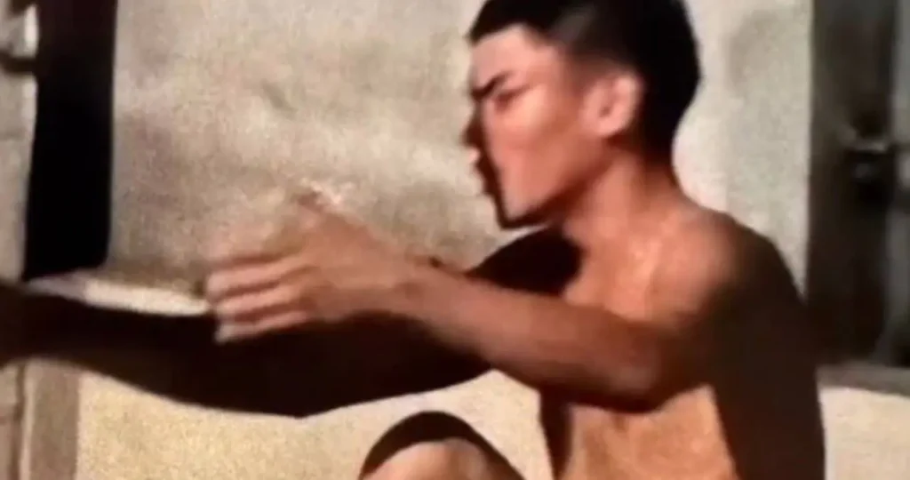 Em uma rua asfaltada e mal iluminada à noite, um homem com camisa escura e bermuda jeans está sentado no chão com as mãos algemadas para trás. Ele é vigiado por dois policiais militares de Alagoas, que vestem fardamento cinza, coletes balísticos e portam armas longas. Ao fundo, as luzes de uma viatura e de residências próximas.