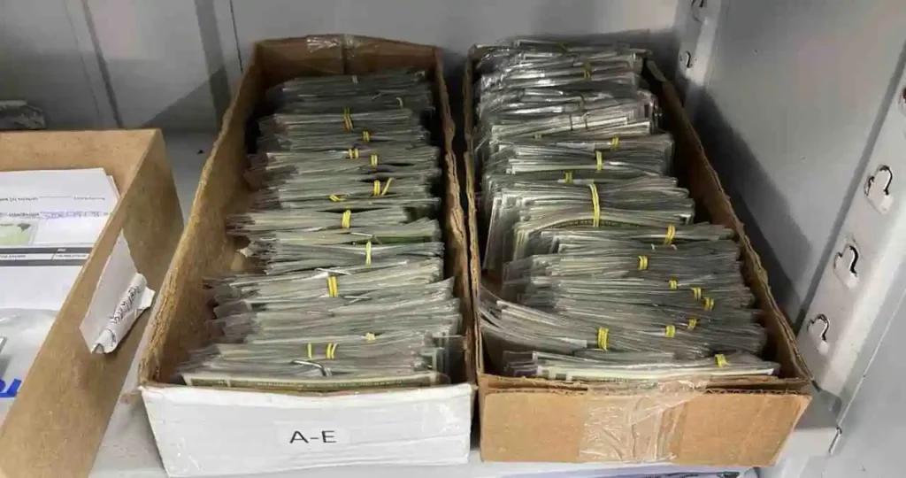 Duas caixas de papelão abertas, posicionadas lado a lado em uma prateleira cinza, estão repletas de maços de documentos de identidade (CIN) plastificados e organizados verticalmente. Cada maço está preso por um elástico amarelo. A caixa da esquerda possui uma etiqueta branca com a inscrição "A-E".