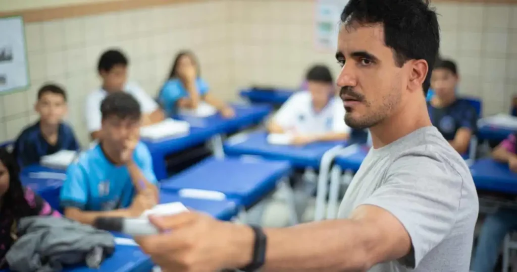 Em uma sala de aula, um professor jovem, de pele parda e barba curta, veste uma camiseta cinza e segura um pincel atômico preto enquanto olha seriamente para a frente. Ao fundo, estudantes em uniformes azuis estão sentados em suas bancas, com cadernos abertos.
