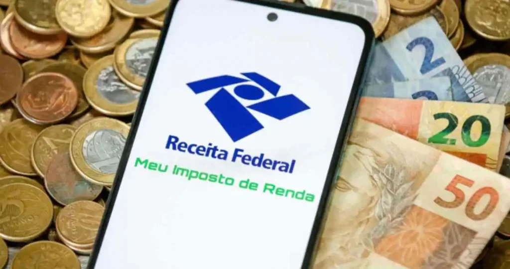 Um smartphone branco está posicionado sobre um fundo repleto de moedas de Real de diversos valores. A tela do aparelho exibe o logotipo da Receita Federal e o texto "Meu Imposto de Renda". À direita do celular, notas de 2, 20 e 50 Reais aparecem parcialmente.