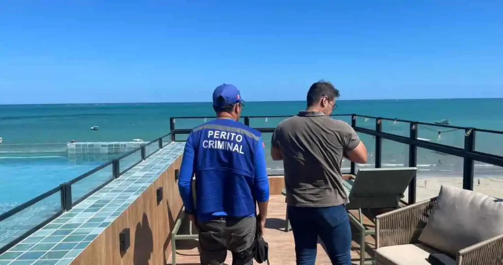 Em um terraço com vista para o mar azul turquesa de Maragogi, dois homens estão de costas observando a área da piscina. O homem à esquerda veste uma camiseta azul com a inscrição "PERITO CRIMINAL" em letras brancas. O homem à direita veste uma camiseta polo marrom e calça jeans. O local possui piso amadeirado, cadeiras de sol e um guarda-corpo metálico com varal de luzes