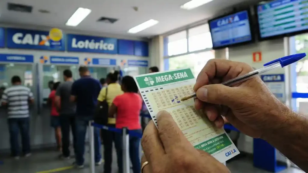Mãos seguram um volante da Mega-Sena e uma caneta dentro de uma lotérica da Caixa, com fila de apostadores ao fundo e painéis de atendimento na parede.
