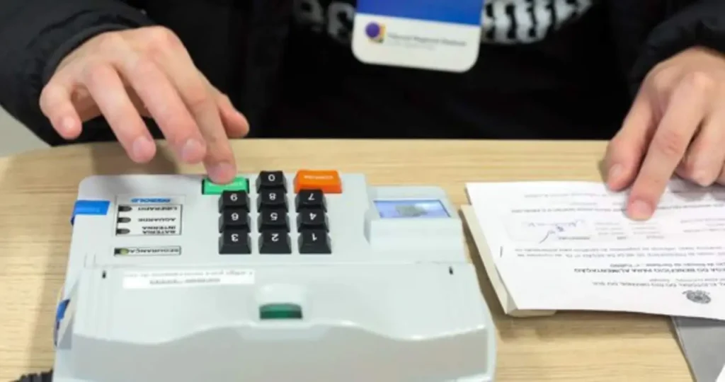 Em um fundo branco, uma urna eletrônica modelo 2020 está inclinada à direita. O teclado numérico em braile e os botões "Branco", "Corrige" e "Confirma" são visíveis. À esquerda, em destaque, o selo circular da Justiça Eleitoral Brasileira. No topo, o texto em letras azuis e pretas: "QUER SER MESÁRIO VOLUNTÁRIO?".