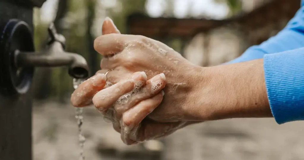 Close-up das mãos de uma pessoa sendo lavadas com sabão sob uma torneira de metal antiga em ambiente externo. A espuma branca cobre os dedos; ao fundo, é possível ver árvores e um ambiente de jardim levemente desfocado.