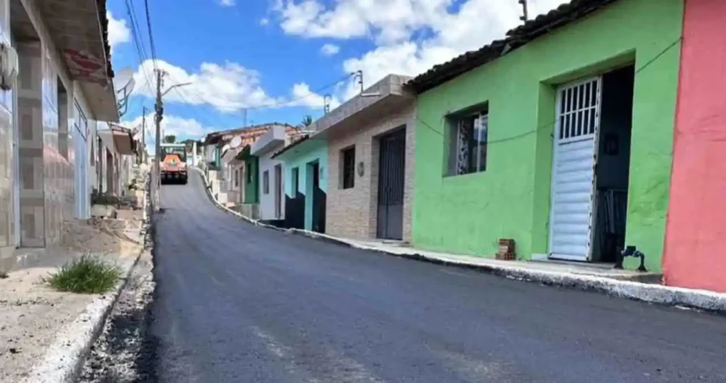 Uma pavimentadora de asfalto amarela, com a inscrição "VÖGELE" em preto, opera em uma rua urbana. O asfalto novo, de cor preta intensa, acaba de ser aplicado. Ao fundo, casas simples com fachadas coloridas e fiação elétrica aérea. O dia está ensolarado e o céu está azul.