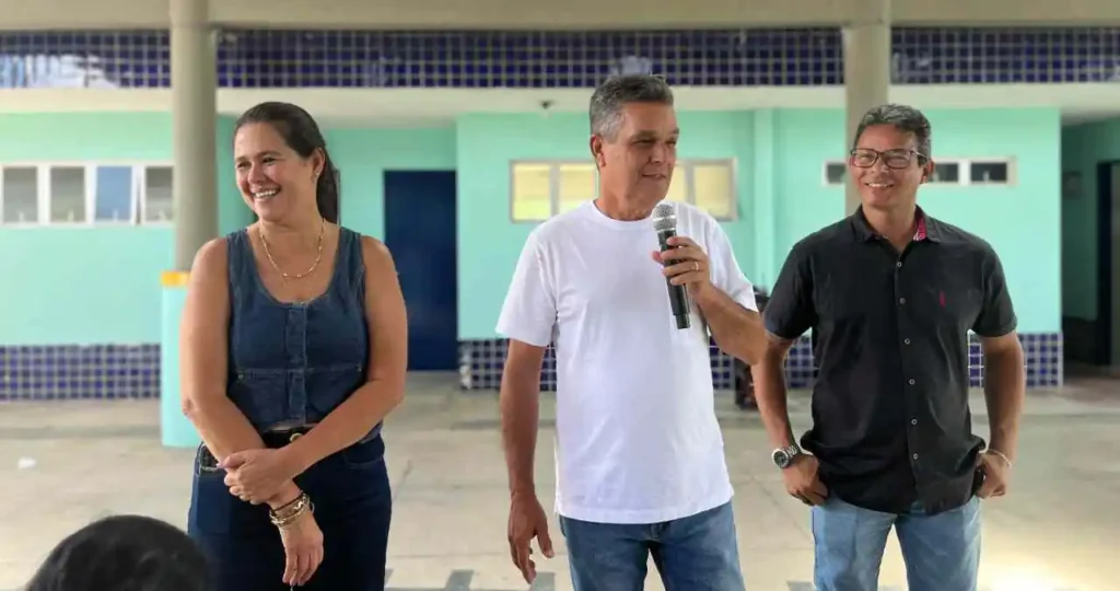 Em um pátio escolar coberto com telhas de barro e estrutura de vigas de concreto, três pessoas estão em pé. No centro, o prefeito Neno Freitas, de camiseta branca e jeans, fala ao microfone. À sua esquerda, uma mulher de colete jeans sorri, e à sua direita, o secretário Ednaldo Firmino, de camisa preta, observa. O fundo mostra salas de aula com paredes verdes e detalhes em azulejos azuis.