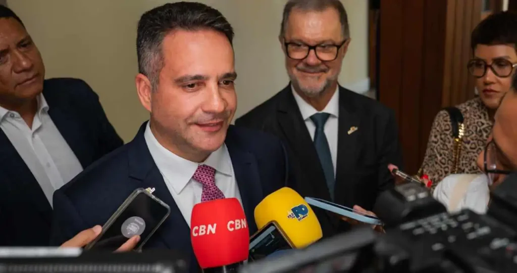 O governador de Alagoas, Paulo Dantas, vestindo um terno azul marinho e gravata rosa, concede entrevista cercado por microfones de diversas emissoras, incluindo CBN e TV Pajuçara. Ao fundo, assessores e câmeras de TV registram o momento em um ambiente interno bem iluminado.