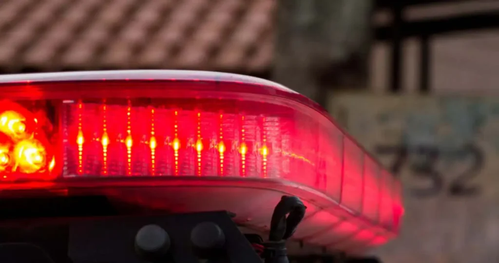 Close-up em ângulo lateral do giroflex de uma viatura policial. A barra de luzes vermelha está acesa e brilhando intensamente. Ao fundo, de forma desfocada, observa-se o telhado de uma residência e o número "732" pintado em uma parede.