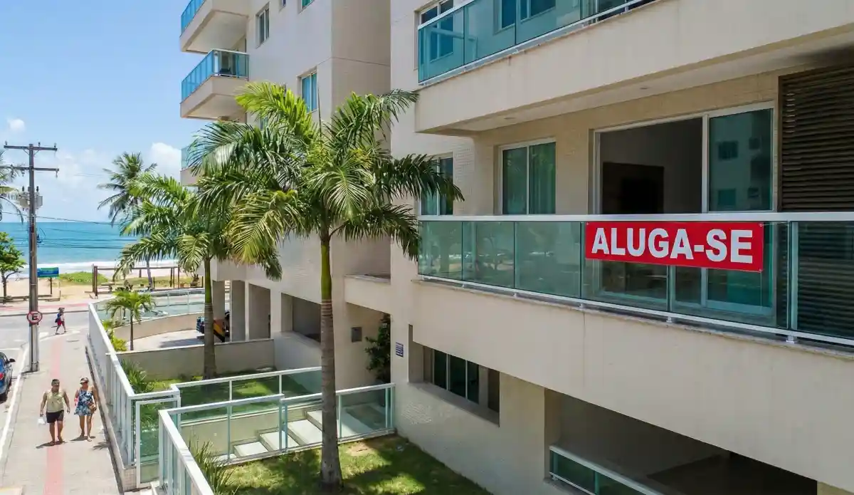 Prédio residencial à beira-mar com placa “aluga-se” na varanda, com coqueiros e orla ao fundo em Alagoas