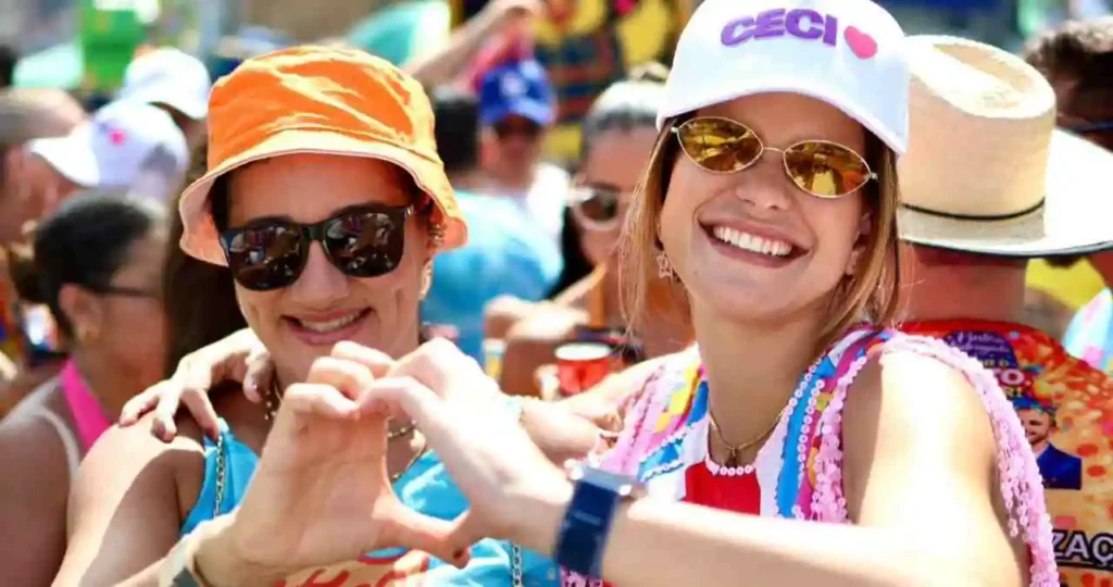 Duas mulheres sorridentes posam juntas em meio à multidão do bloco Pinto da Madrugada. À esquerda, uma mulher usa chapéu bucket laranja e óculos escuros, fazendo o sinal de coração com as mãos. À direita, a prefeita Ceci, usando um boné branco com seu nome e óculos de sol, completa o gesto de coração. Ambas vestem abadás coloridos com temas carnavalescos e feministas.