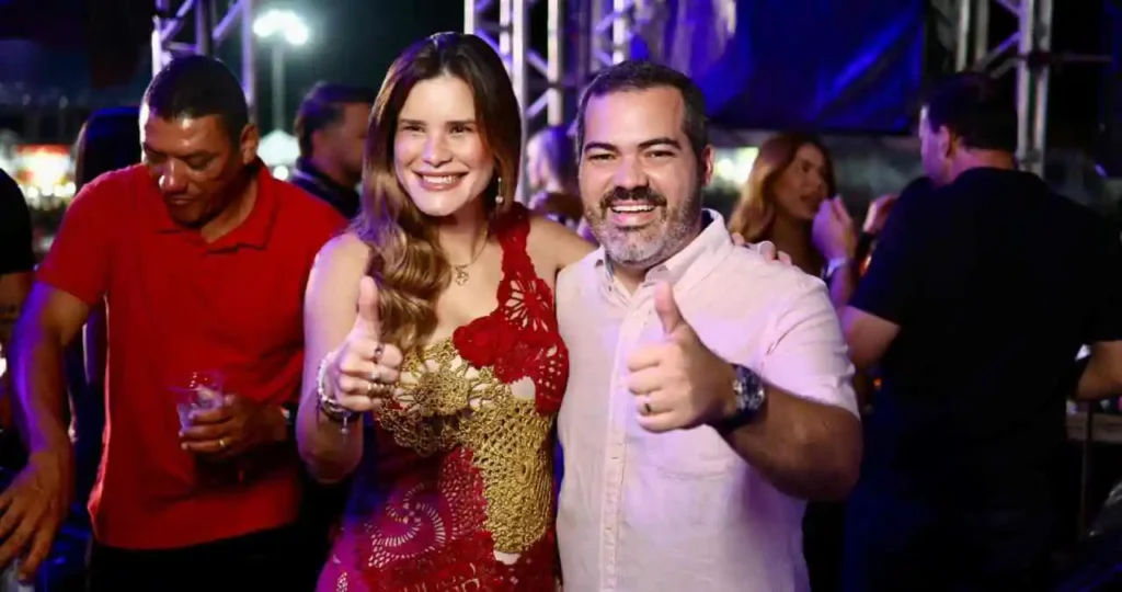 A prefeita de Atalaia, Ceci, vestindo um vestido vermelho rendado, e o vice-prefeito Nicollas, de camisa polo rosa clara, sorriem e fazem sinal de "positivo" com os polegares em um evento noturno. Ao fundo, luzes de palco e pessoas em um ambiente de festa.